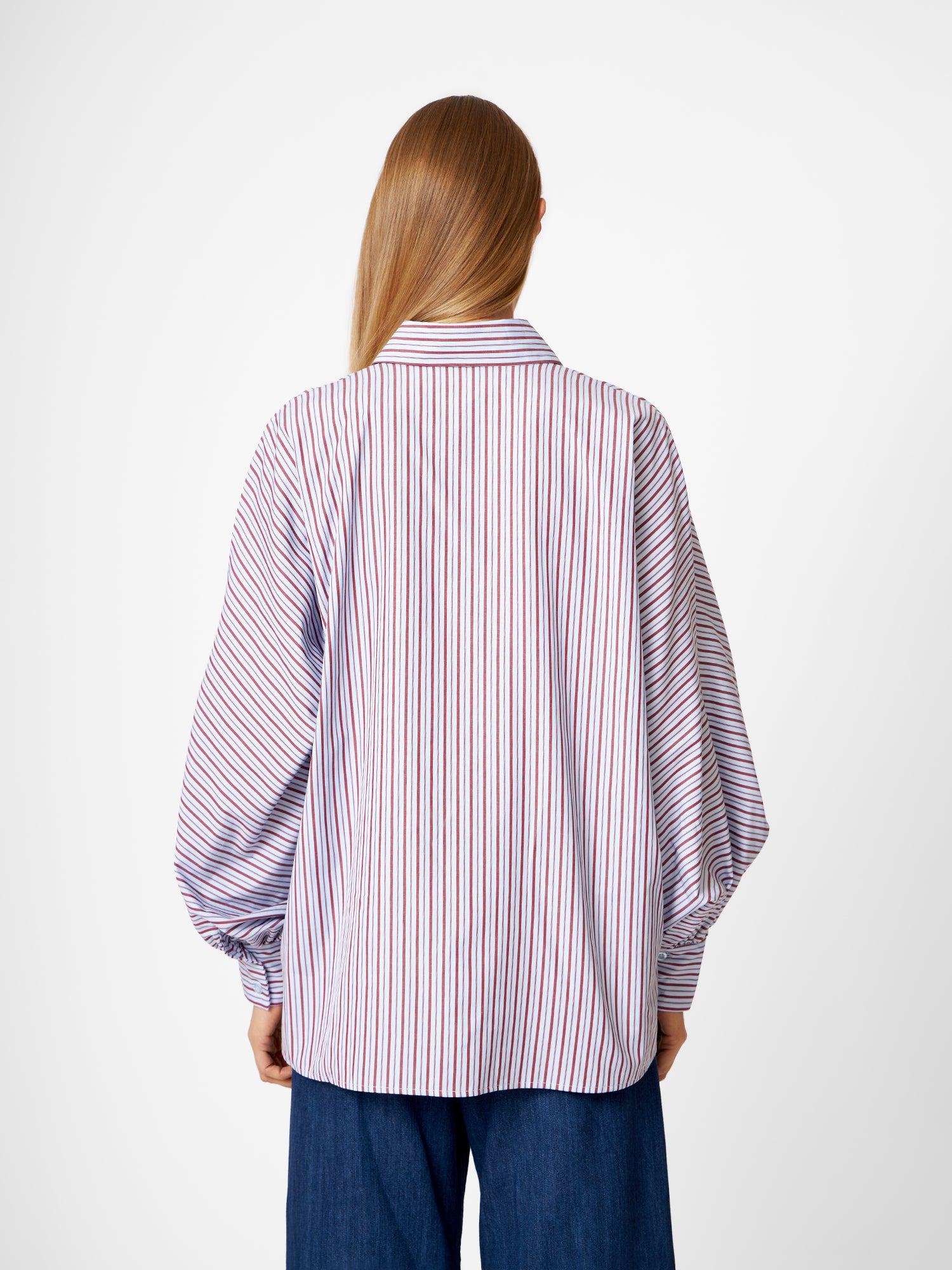Eine Person mit langen glatten Haaren, von hinten gesehen, trägt die Bluse Antonia Blue Burgundy Stripe und eine dunkelblaue Hose. Der Hintergrund ist einfarbig weiß.