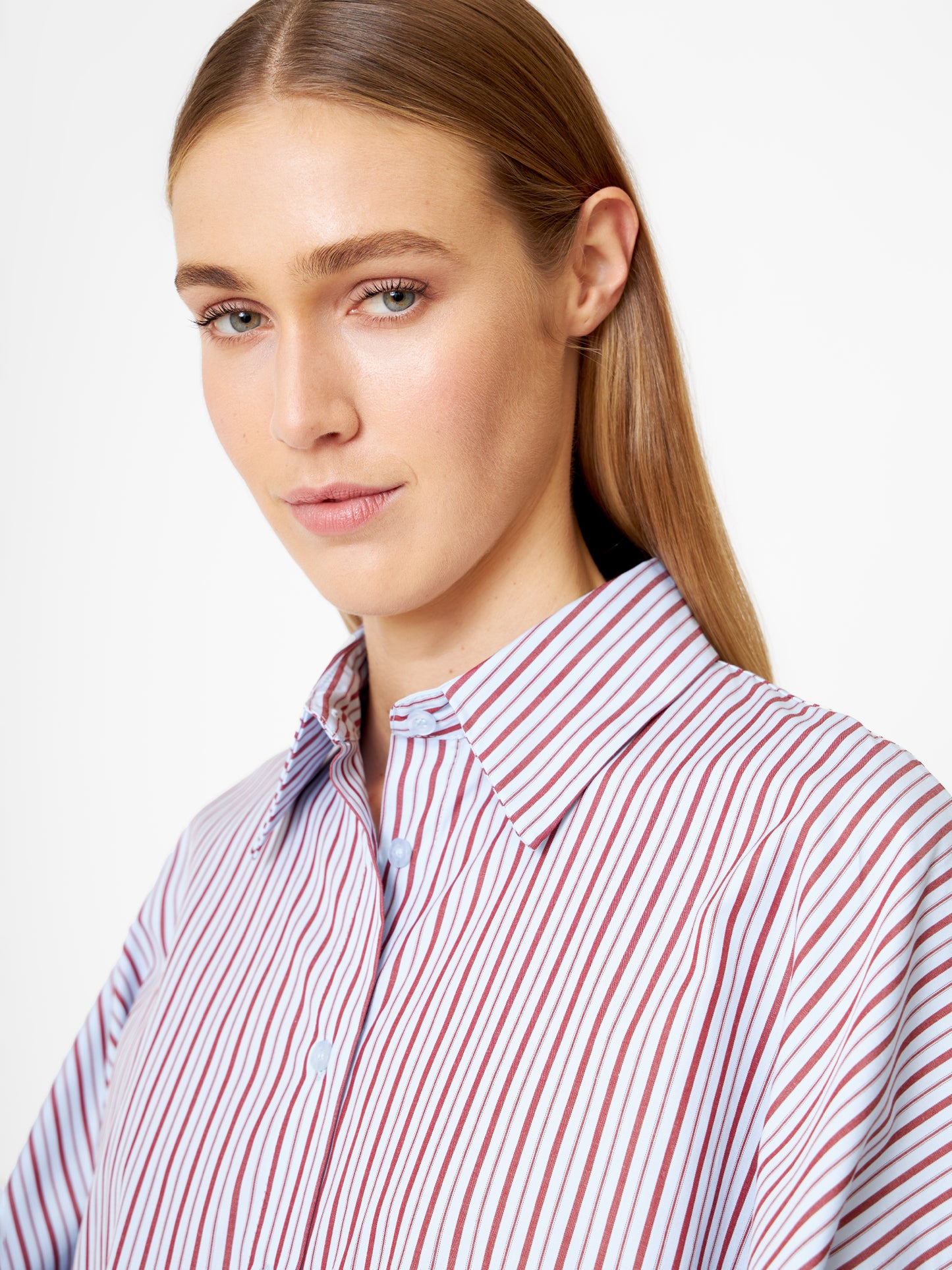 Eine Frau mit glattem, hellbraunem Haar trägt die Bluse Antonia Blue Burgundy Stripe. Sie blickt leicht zur Seite und hat einen neutralen Ausdruck vor einem schlichten Hintergrund.
