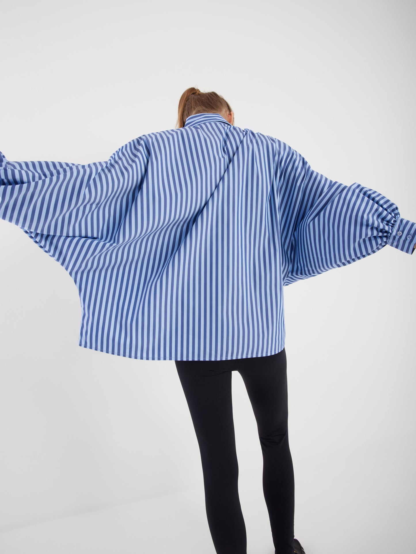 Eine Person steht mit ausgestreckten Armen, trägt die Bluse Antonia light mit dunkelblauen Streifen und schwarzer Hose und blickt von der Kamera weg auf einen schlichten weißen Hintergrund.
