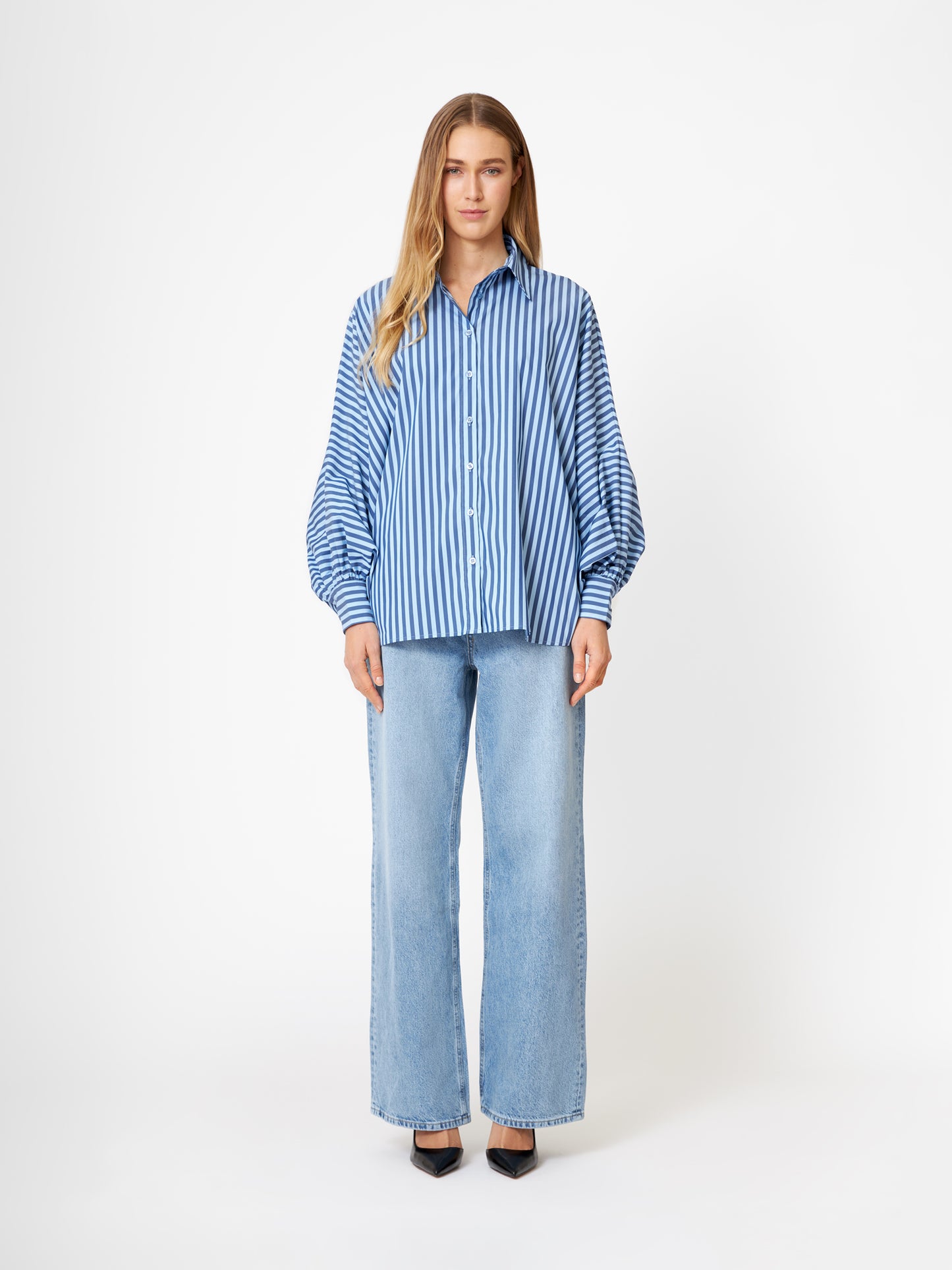 Eine Frau blickt nach vorne und trägt die Bluse Antonia light, dark blue stripe - eine langärmelige, blau-weiß gestreifte Bluse - mit hellblauen, weit geschnittenen Jeans und schwarzen spitzen Schuhen vor einem schlichten weißen Hintergrund.