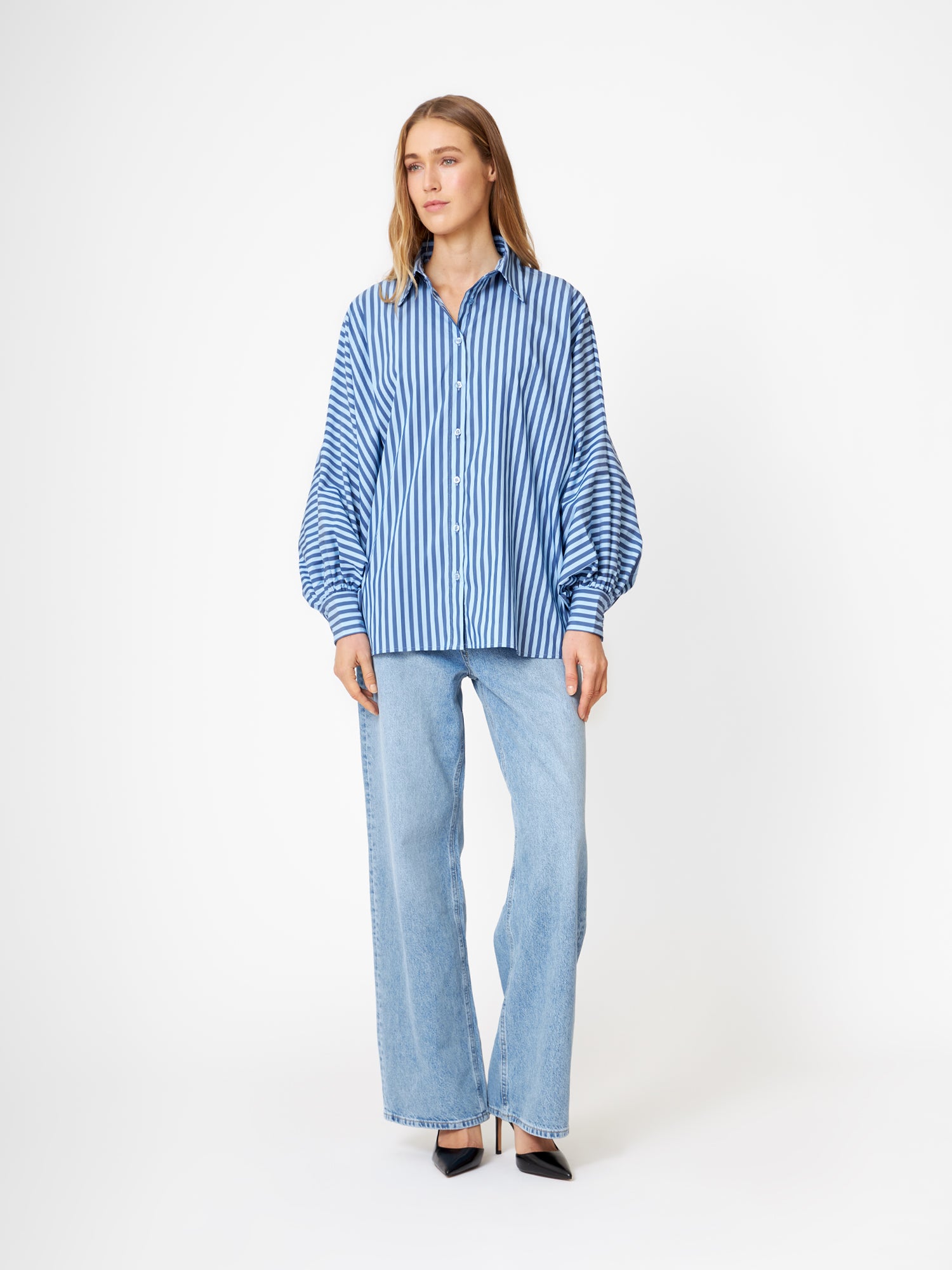 Eine Frau steht vor einem weißen Hintergrund und trägt die Bluse Antonia light mit dunkelblauen Streifen und Ballonärmeln, gepaart mit hellblauen Jeans mit weitem Bein und schwarzen spitzen Absätzen.