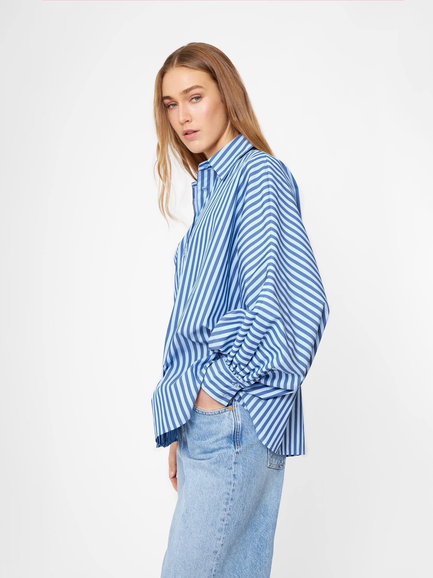 Eine Frau mit langem, hellbraunem Haar trägt die Bluse Antonia light in dunkelblauen Streifen und hellblauen Jeans und steht seitlich vor einem schlichten weißen Hintergrund.