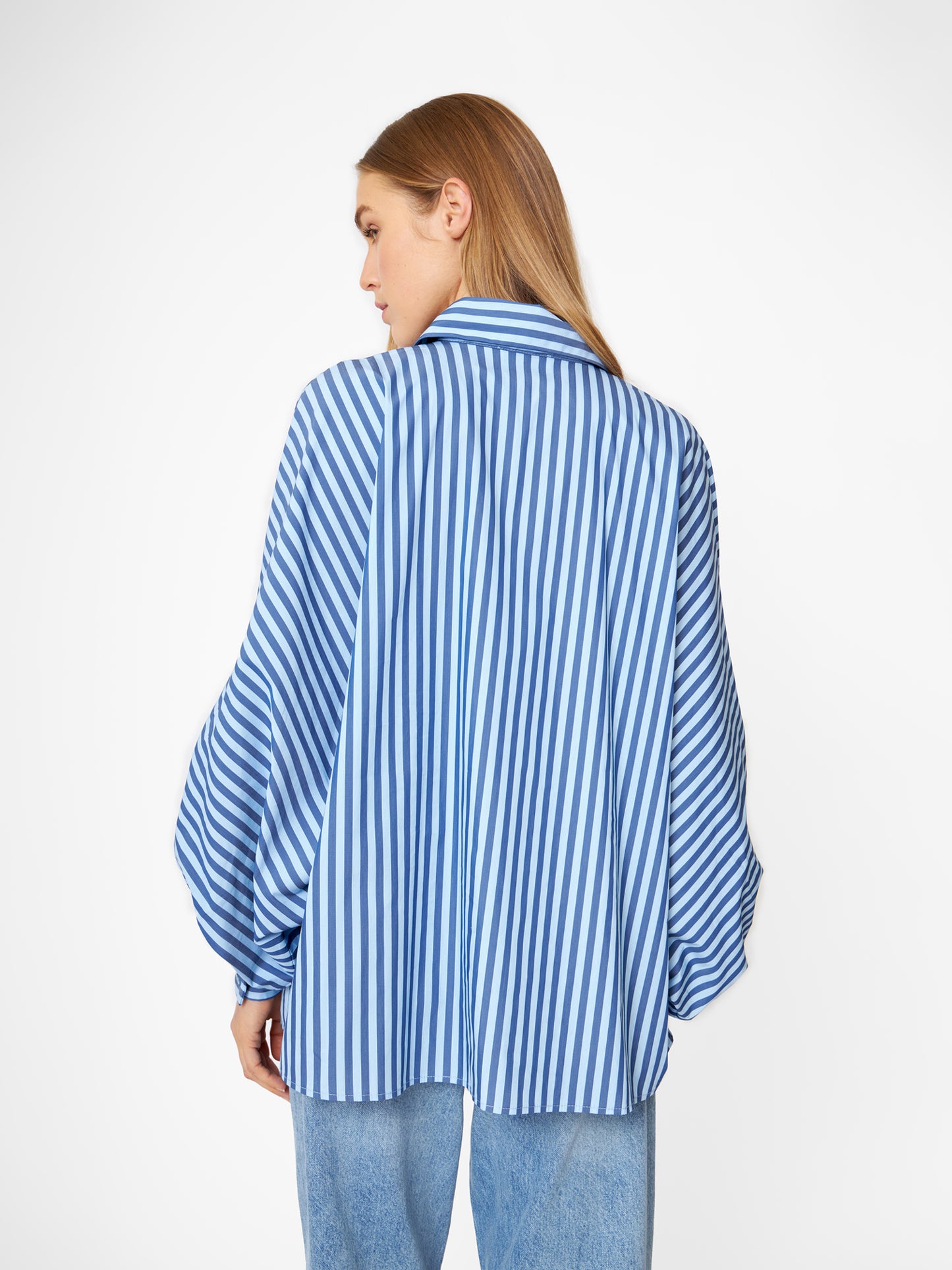 Eine Frau mit langen blonden Haaren trägt die Bluse Antonia light mit dunkelblauen Streifen und hellblauen Jeans und steht mit dem Rücken zur Kamera vor einem schlichten weißen Hintergrund.