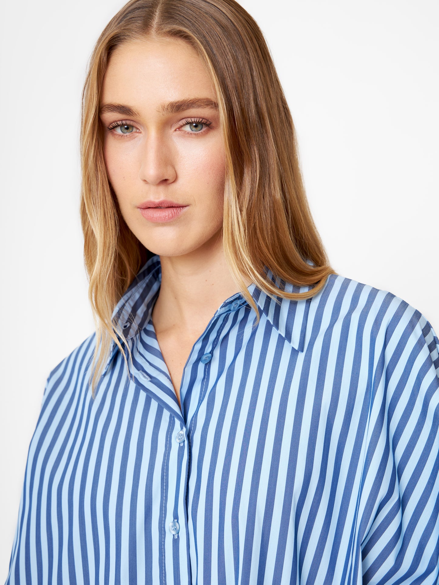 Eine Frau mit langen blonden Haaren trägt die Bluse Antonia hell, dunkelblau gestreift, gestylt als Oversize Damen Hemd. Sie blickt mit neutralem Gesichtsausdruck vor einem schlichten weißen Hintergrund in die Kamera.