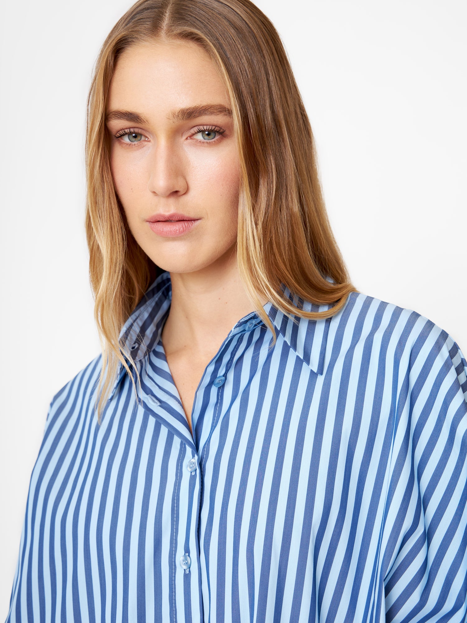 Eine Frau mit langen blonden Haaren trägt die Bluse Antonia hell, dunkelblau gestreift, gestylt als Oversize Damen Hemd. Sie blickt mit neutralem Gesichtsausdruck vor einem schlichten weißen Hintergrund in die Kamera.