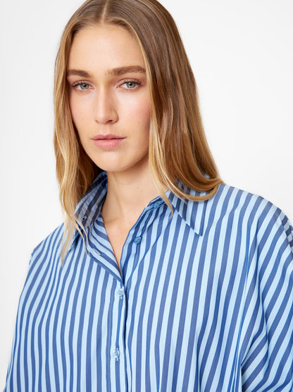 Eine Frau mit langen blonden Haaren trägt die Bluse Antonia hell, dunkelblau gestreift, gestylt als Oversize Damen Hemd. Sie blickt mit neutralem Gesichtsausdruck vor einem schlichten weißen Hintergrund in die Kamera.