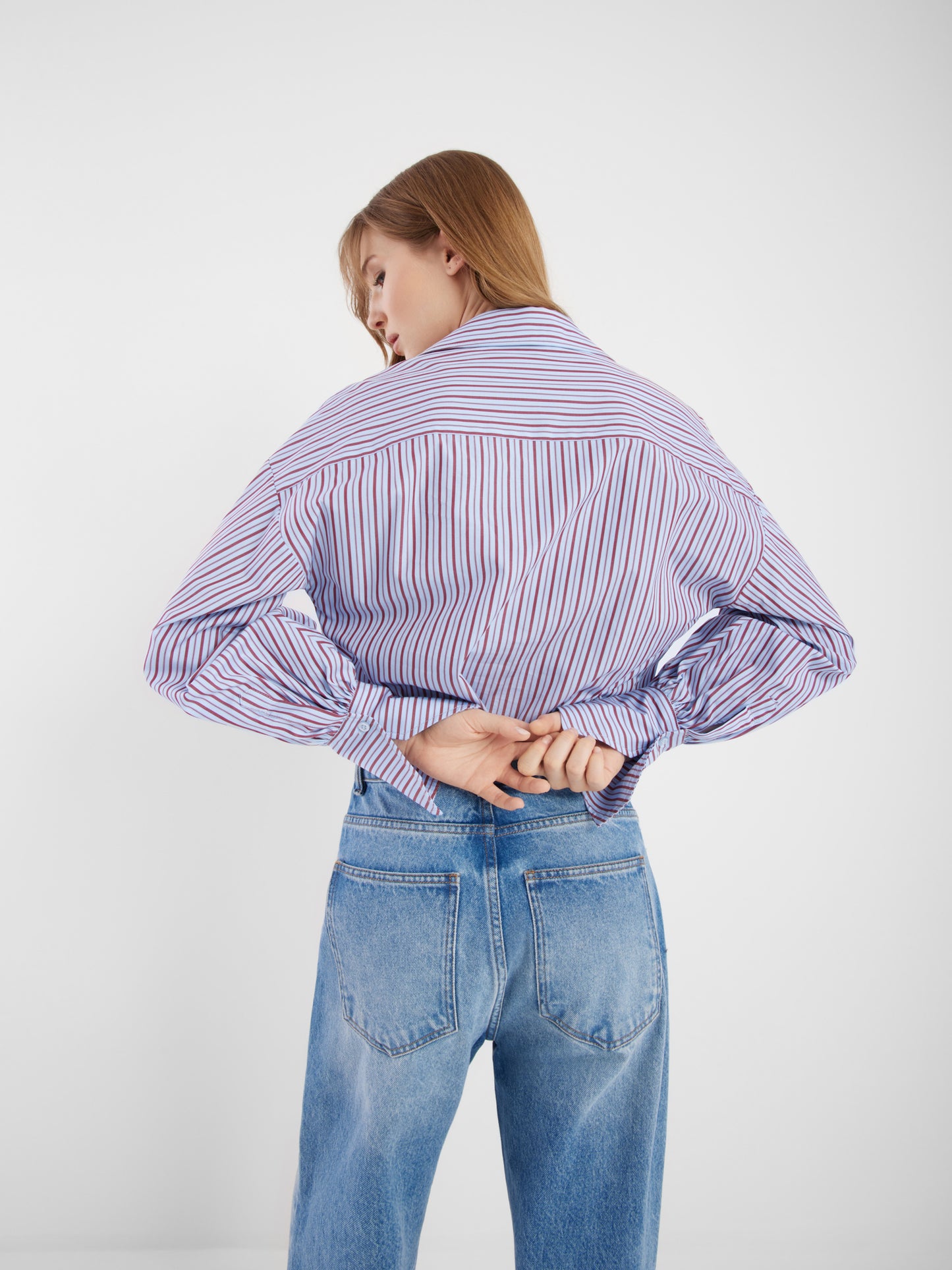 Eine Person mit glattem, hellbraunem Haar steht mit dem Rücken zur Kamera, trägt die Bluse Claire Blue Burgundy Stripe und hellblaue Jeans, die Hände hinter dem Rücken verschränkt.
