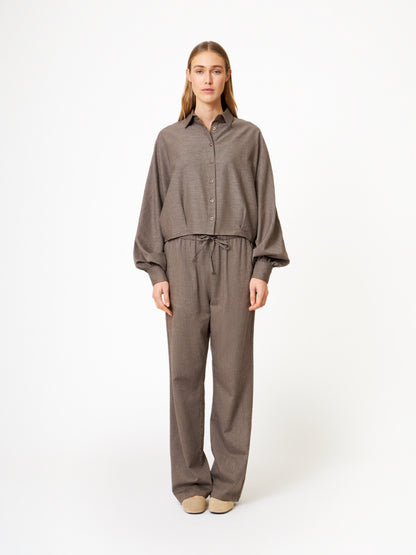 Eine Frau in einem lockeren braunen Button-up-Hemd und einer Hose Flanell Taupe mit Kordelzug in der Taille, dazu beigefarbene Schuhe, präsentiert winterlichen Streetstyle vor einem schlichten weißen Hintergrund.