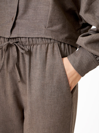 Eine Person trägt eine locker sitzende, langärmelige taupefarbene Flanell-Hose (Pants Flanell Taupe) mit elastischem Bund und Kordelzug, die rechte Hand in der Tasche - ideal für einen gemütlichen Winter-Streetstyle - vor einem schlichten weißen Hintergrund.