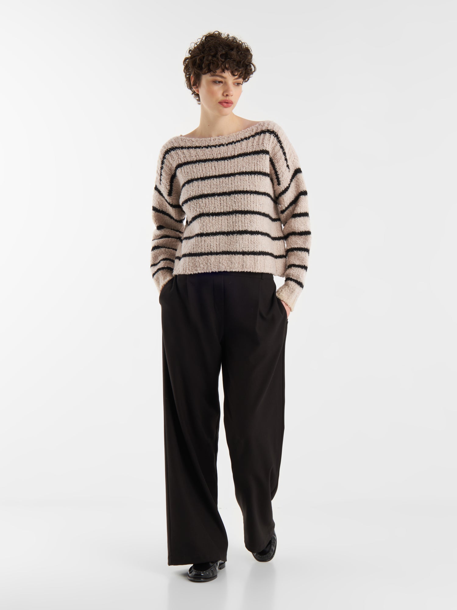 Eine Person mit kurzen lockigen Haaren und einer schwarzen Hose mit weitem Bein posiert mit den Händen in den Taschen vor einem schlichten weißen Hintergrund und trägt den Pullover Petite Sailor Black Stripe.