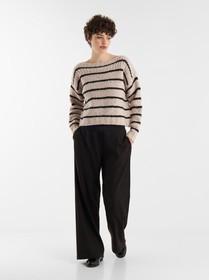 Eine Person mit kurzen lockigen Haaren und einer schwarzen Hose mit weitem Bein posiert mit den Händen in den Taschen vor einem schlichten weißen Hintergrund und trägt den Pullover Petite Sailor Black Stripe.