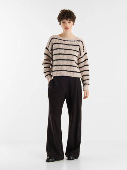 Eine Person mit kurzen lockigen Haaren steht vor einem weißen Hintergrund und trägt den Pullover Petite Sailor Black Stripe, gepaart mit einer weiten schwarzen Hose und schwarzen Schuhen.