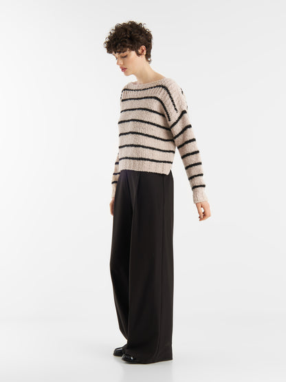 Eine Person mit kurzen lockigen Haaren steht seitlich vor einem einfarbig weißen Hintergrund und trägt den Pullover Petite Sailor Black Stripe mit schwarzen Querstreifen und eine locker sitzende schwarze Hose.