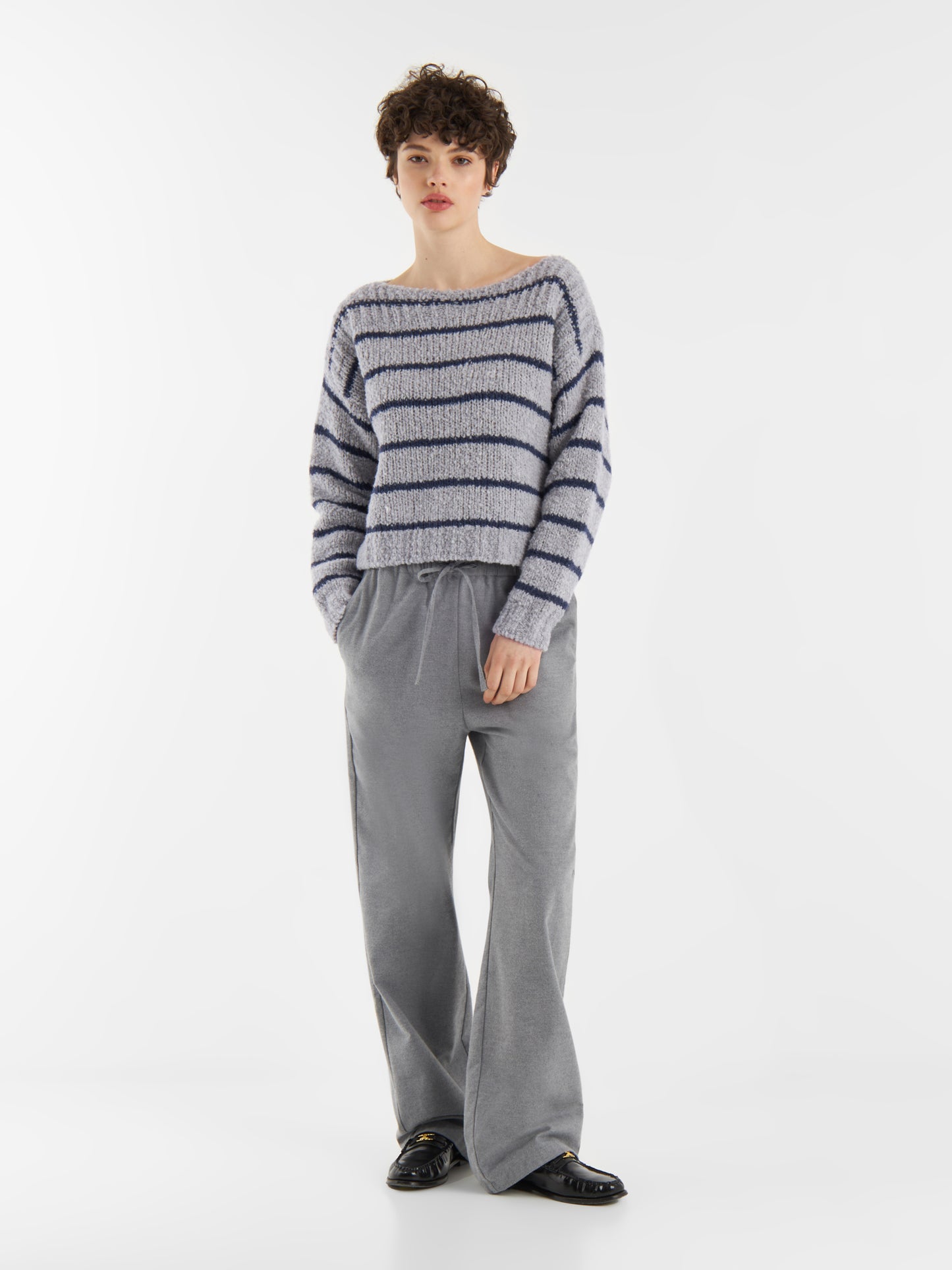 Eine Person mit kurzen lockigen Haaren trägt den blau gestreiften Pullover Petite Sailor, eine graue Hose mit weitem Bein und schwarze Schuhe. Sie steht mit den Händen in den Taschen vor einem schlichten weißen Hintergrund.