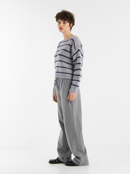 Eine Person mit kurzen lockigen Haaren steht seitlich, trägt den Pullover Petite Sailor blau gestreift und eine lockere graue Hose mit Kordelzug und schwarzen Schuhen. Der kuschelige Pullover hebt sich im Strickpullover Damen Stil von einem schlichten weißen Hintergrund ab.