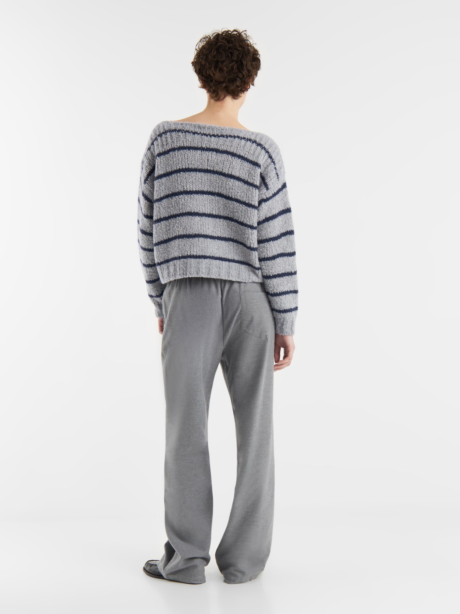 Eine Person blickt von sich weg und trägt den Pullover Petite Sailor blue stripe - ein flauschiges hellgraues Oberteil mit dunkelblauen Streifen - und eine weite graue Hose vor einem schlichten weißen Hintergrund.