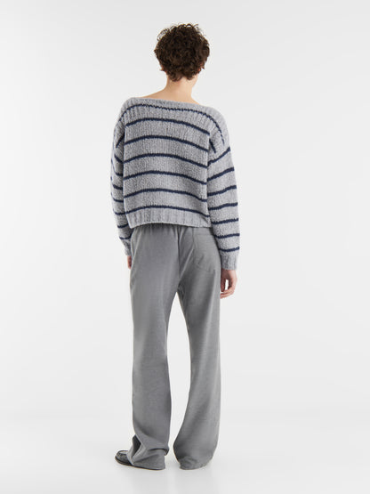 Eine Person blickt von sich weg und trägt den Pullover Petite Sailor blue stripe - ein flauschiges hellgraues Oberteil mit dunkelblauen Streifen - und eine weite graue Hose vor einem schlichten weißen Hintergrund.