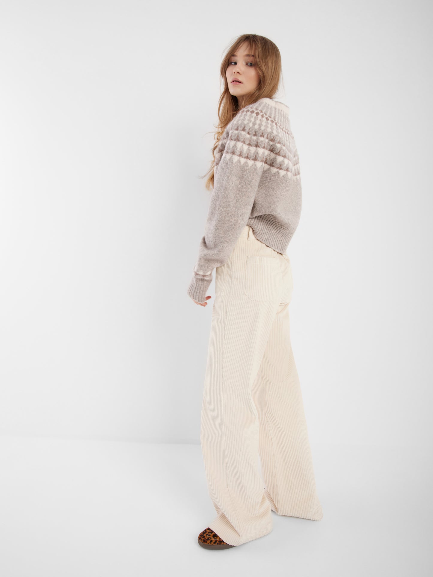Eine Frau mit langem, gewelltem Haar steht seitlich und trägt den grauen Norweger-Pullover und eine hochgezogene cremefarbene Hose mit weitem Bein und braunen Schuhen, die den skandinavischen Stil vor einem schlichten weißen Hintergrund zeigen.