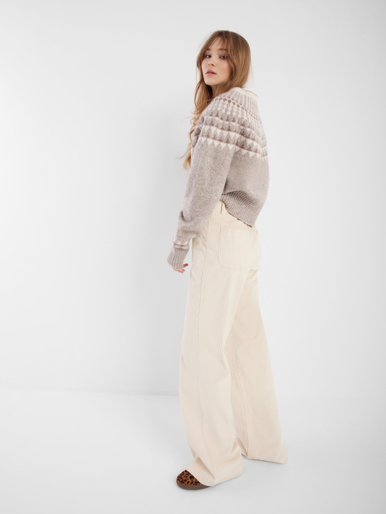Eine Frau mit langem, gewelltem Haar steht seitlich und trägt den grauen Norweger-Pullover und eine hochgezogene cremefarbene Hose mit weitem Bein und braunen Schuhen, die den skandinavischen Stil vor einem schlichten weißen Hintergrund zeigen.