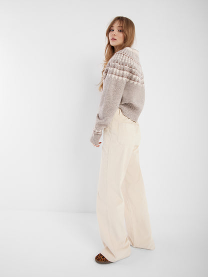 Eine Frau mit langem, gewelltem Haar steht seitlich und trägt den grauen Norweger-Pullover und eine hochgezogene cremefarbene Hose mit weitem Bein und braunen Schuhen, die den skandinavischen Stil vor einem schlichten weißen Hintergrund zeigen.