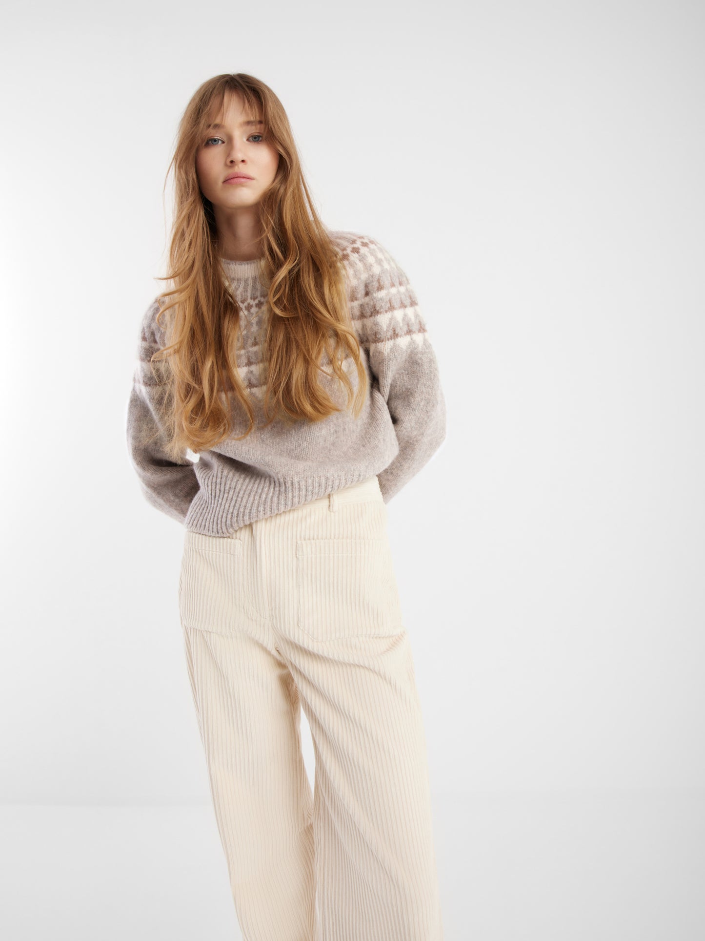 Eine junge Frau mit langem, gewelltem blondem Haar trägt einen hellgrauen Norweger-Pullover und eine hochtaillierte, hellbeige Cordhose und verkörpert damit skandinavischen Stil vor einem schlichten weißen Hintergrund.