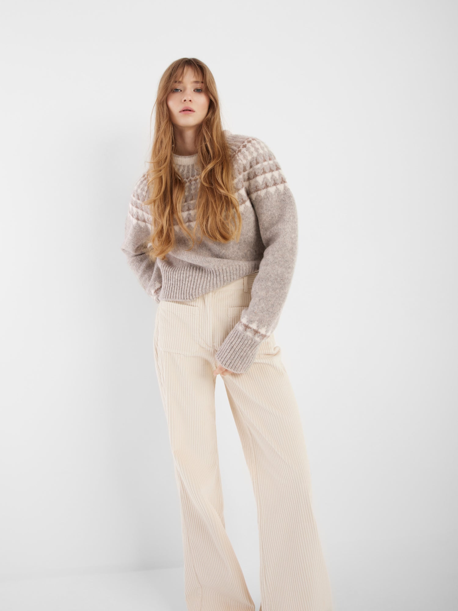Eine Frau mit langem, gewelltem Haar modelliert den grauen Norweger-Pullover und die cremefarbene Hose mit weitem Bein, die den skandinavischen Stil vor einem schlichten weißen Hintergrund verkörpern.