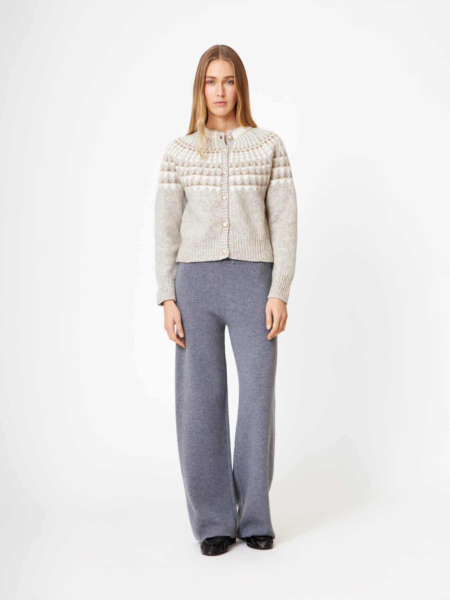 Eine Frau mit glattem, langem Haar und neutralem Gesichtsausdruck steht vor einem schlichten weißen Hintergrund und trägt die gemusterte Jacke Norweger Vol II Greige, eine graue Hose mit weitem Bein und schwarze Schuhe.