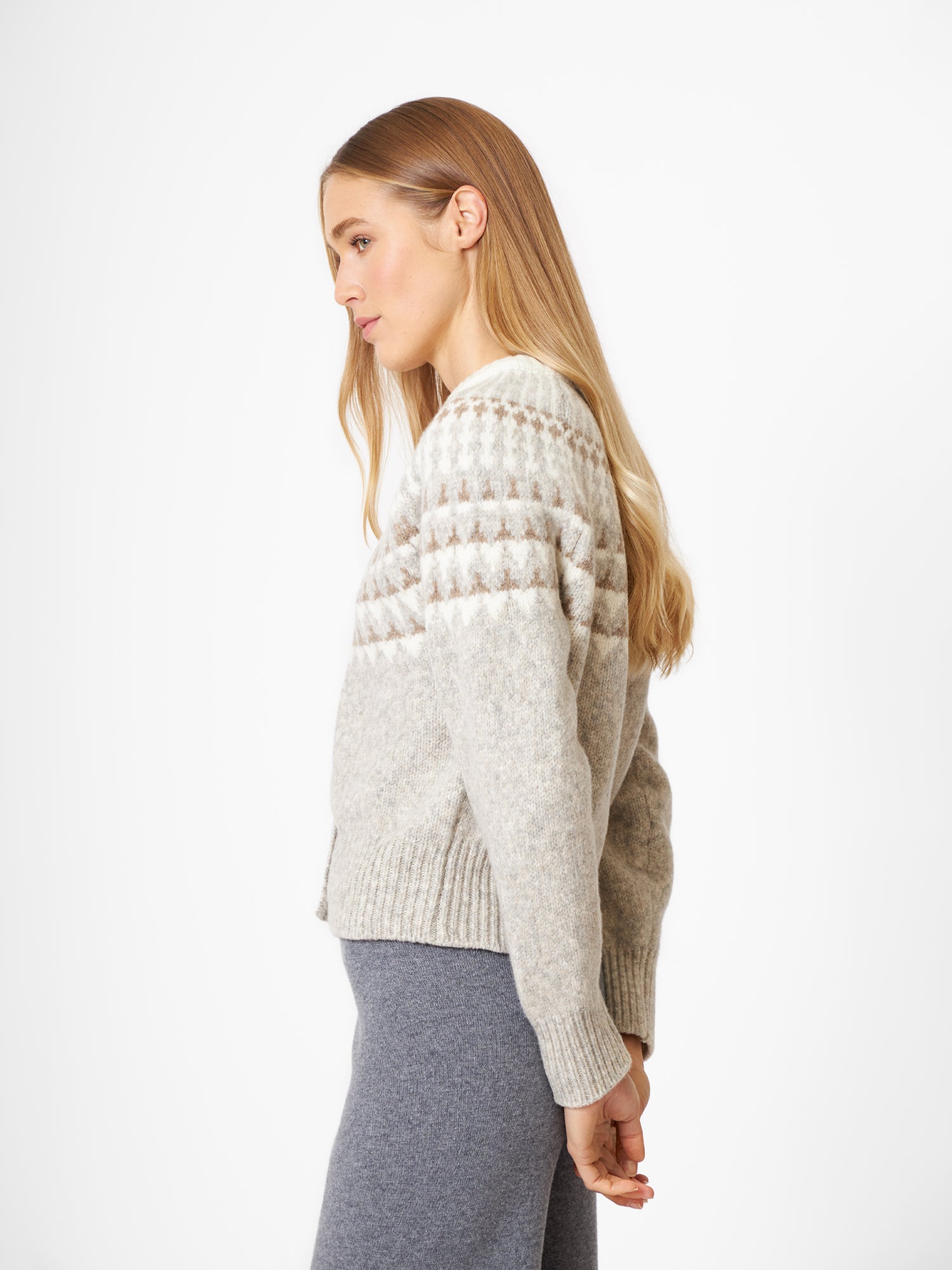 Eine Frau mit langen blonden Haaren steht im Profil vor einem weißen Hintergrund. Sie trägt die Jacke Norweger Vol II Greige und eine graue Hose und hat die Hände hinter dem Rücken.