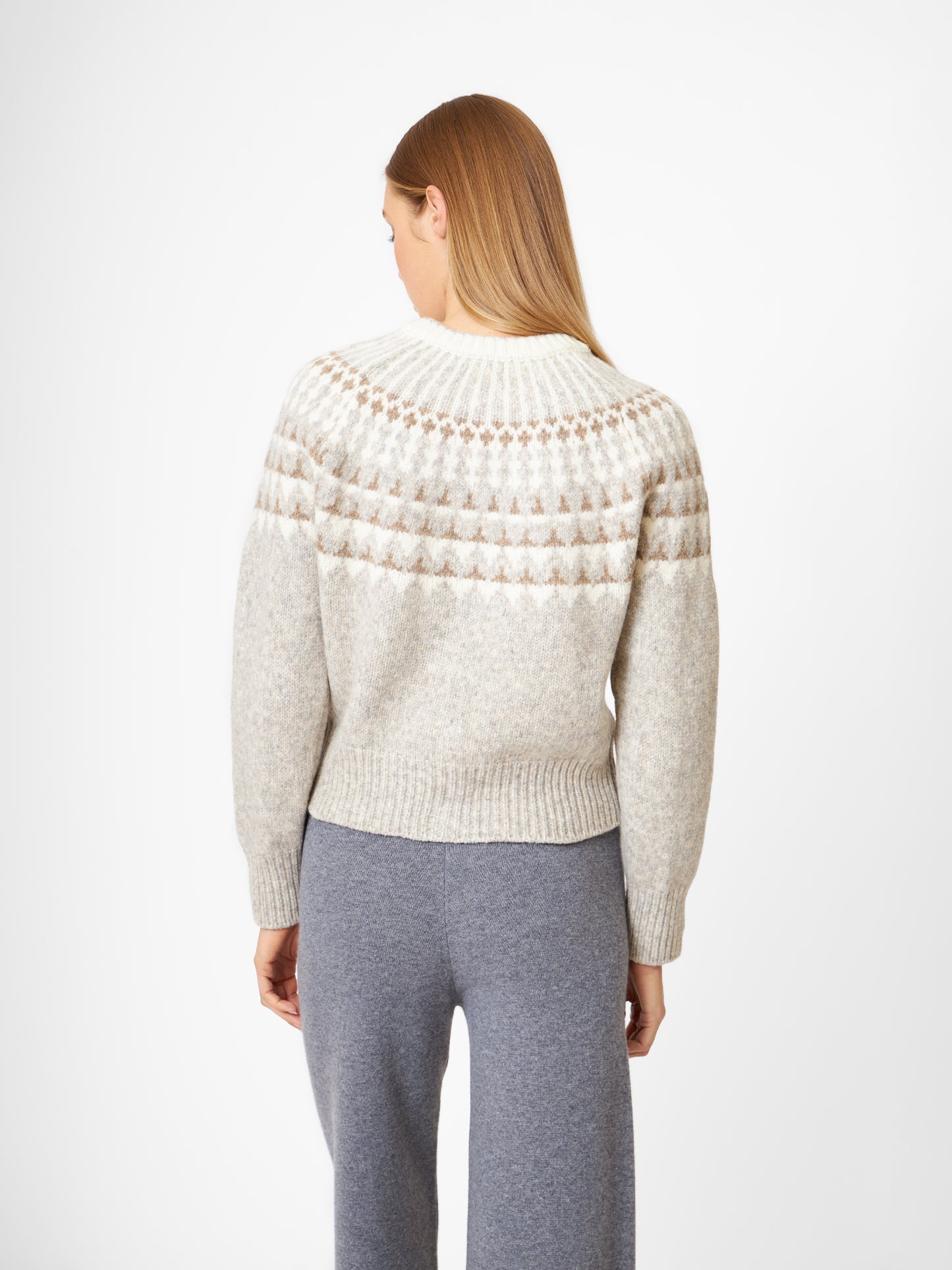 Eine Person mit langem, glattem blondem Haar steht mit dem Rücken zur Kamera und trägt eine hellgraue Jacke Norweger Vol II Greige mit einer gemusterten Passe und einer grauen Hose vor einem schlichten weißen Hintergrund.