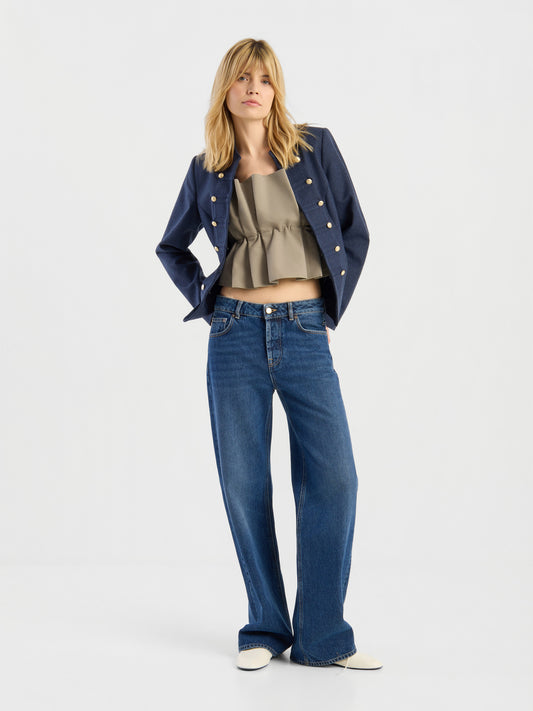 Vor einem weißen Hintergrund steht eine Frau und präsentiert den „Casual Meets Chic“-Look, bestehend aus einem marineblauen Blazer, einem olivgrünen, gerüschten Crop-Top, einer blau-weit geschnittenen Jeans und weißen Schuhen. Mit hinter dem Rücken verschränkten Händen blickt sie leicht zur Seite.