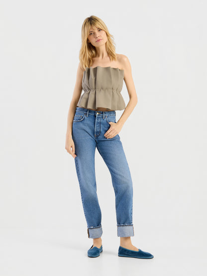 Eine Frau mit blonden Haaren trägt ein olivfarbenes Plissee-Oberteil, eine blaue Jeans mit Umschlag und blaue flache Schuhe und steht vor einem schlichten weißen Hintergrund.