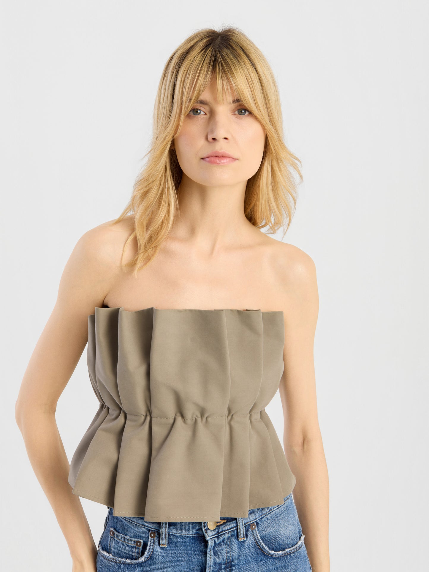 Eine Frau mit blonden Haaren trägt das „Pleat Top Olive“ und eine blaue Jeans und steht vor einem schlichten hellgrauen Hintergrund.