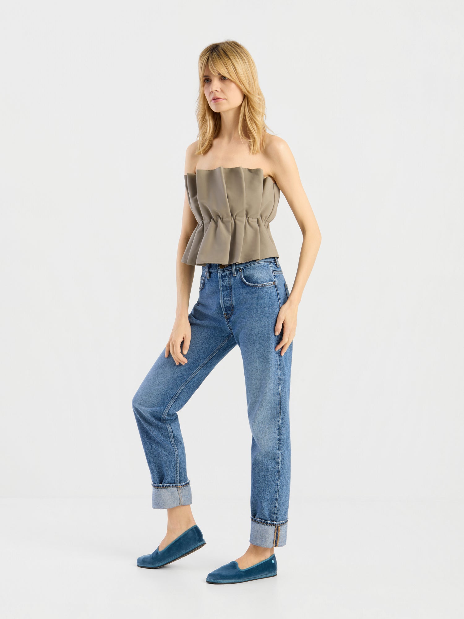 Eine blonde Frau steht vor einem weißen Hintergrund und trägt das „Pleat Top Olive“, eine blaue Jeans mit Umschlag und türkisfarbene Slipper. Sie hat eine entspannte Körperhaltung und steckt eine Hand in die Tasche.