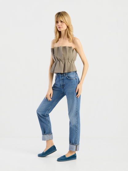 Eine blonde Frau steht vor einem weißen Hintergrund und trägt das „Pleat Top Olive“, eine blaue Jeans mit Umschlag und türkisfarbene Slipper. Sie hat eine entspannte Körperhaltung und steckt eine Hand in die Tasche.