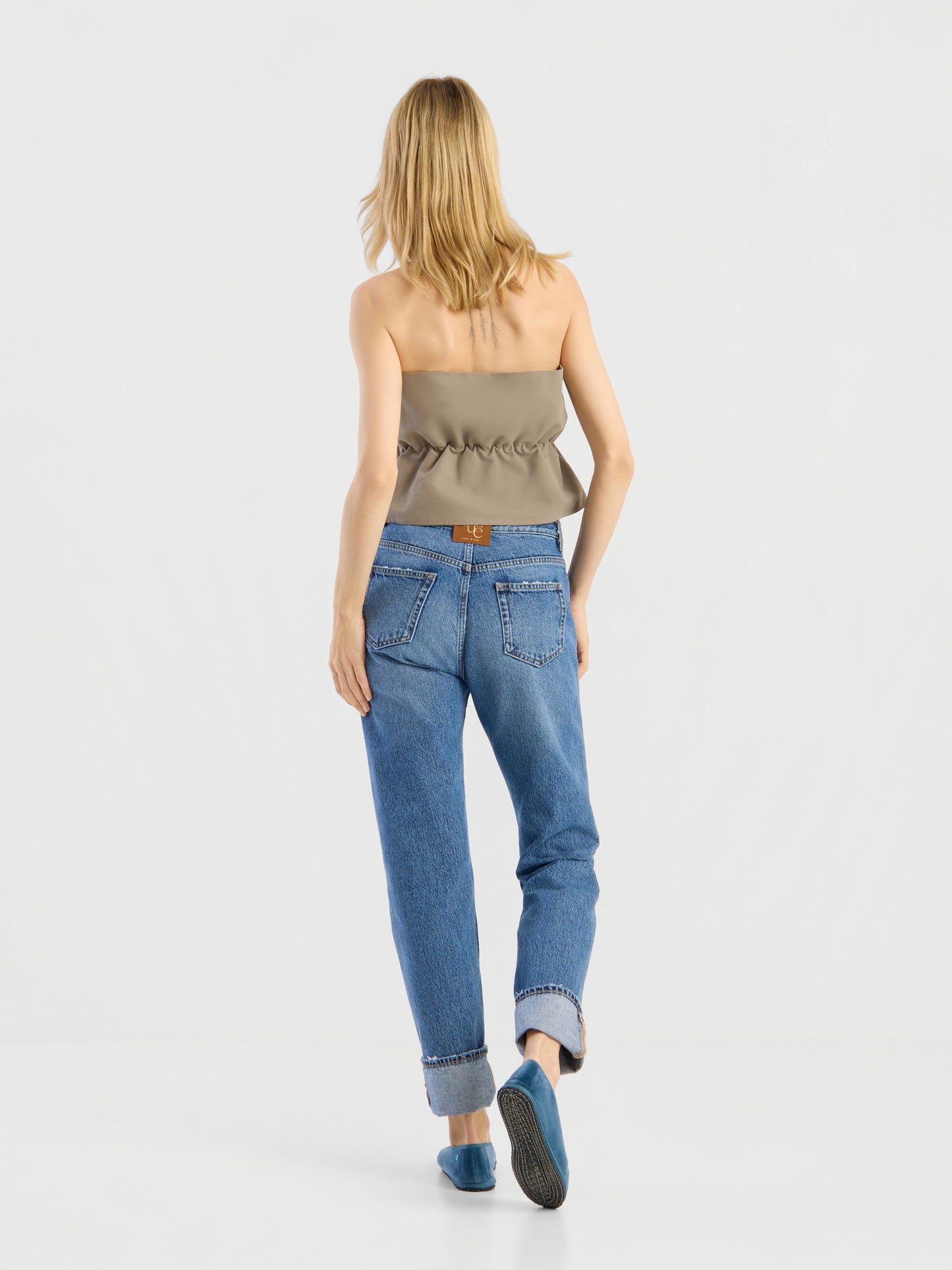 Eine Frau mit blonden Haaren, von hinten gesehen, trägt das „Pleat Top Olive“, eine blaue Jeans mit Umschlag, sichtbaren Gesäßtaschen und einem braunen Aufnäher sowie blaue flache Schuhe und geht vor einem schlichten weißen Hintergrund.