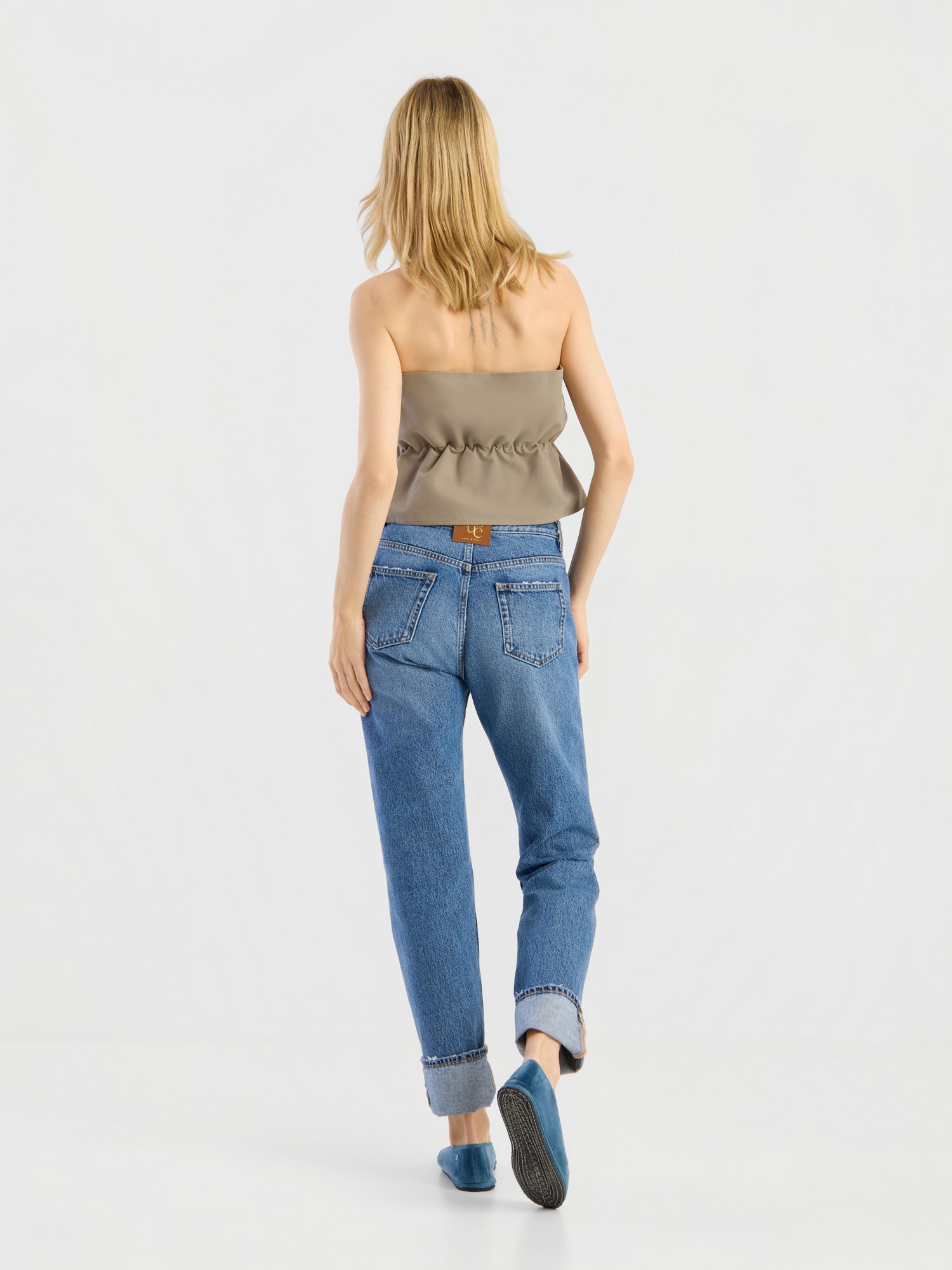 Eine Frau mit blonden Haaren, von hinten gesehen, trägt das „Pleat Top Olive“, eine blaue Jeans mit Umschlag, sichtbaren Gesäßtaschen und einem braunen Aufnäher sowie blaue flache Schuhe und geht vor einem schlichten weißen Hintergrund.