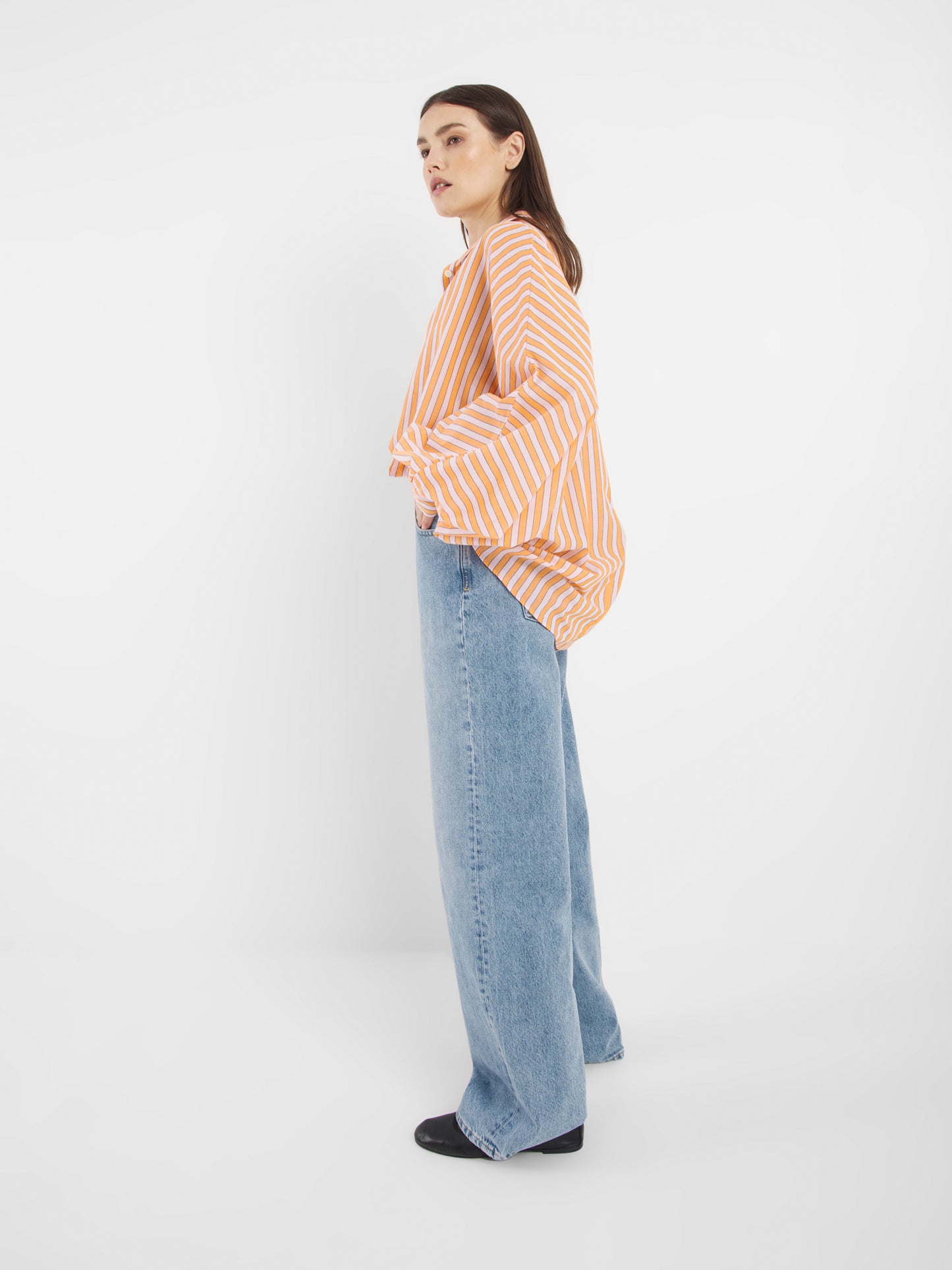 Eine Person steht seitlich vor einem weißen Hintergrund und trägt die Bluse Antonia Rose Coral, gepaart mit einer weit geschnittenen Bluejeans und schwarzen Schuhen.