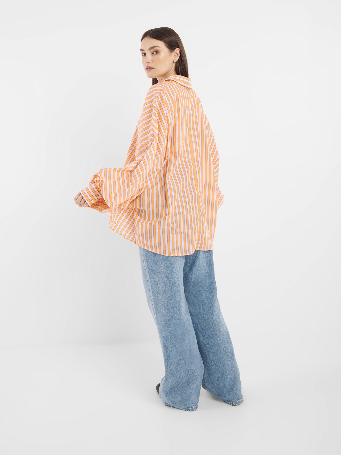 Jemand, der die Bluse Antonia Rose Coral, ein übergroßes orange-weiß gestreiftes Hemd und eine weit geschnittene Bluejeans trägt, steht vor einem schlichten weißen Hintergrund und blickt über seine Schulter zurück.