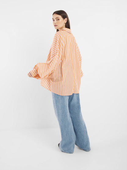 Jemand, der die Bluse Antonia Rose Coral, ein übergroßes orange-weiß gestreiftes Hemd und eine weit geschnittene Bluejeans trägt, steht vor einem schlichten weißen Hintergrund und blickt über seine Schulter zurück.