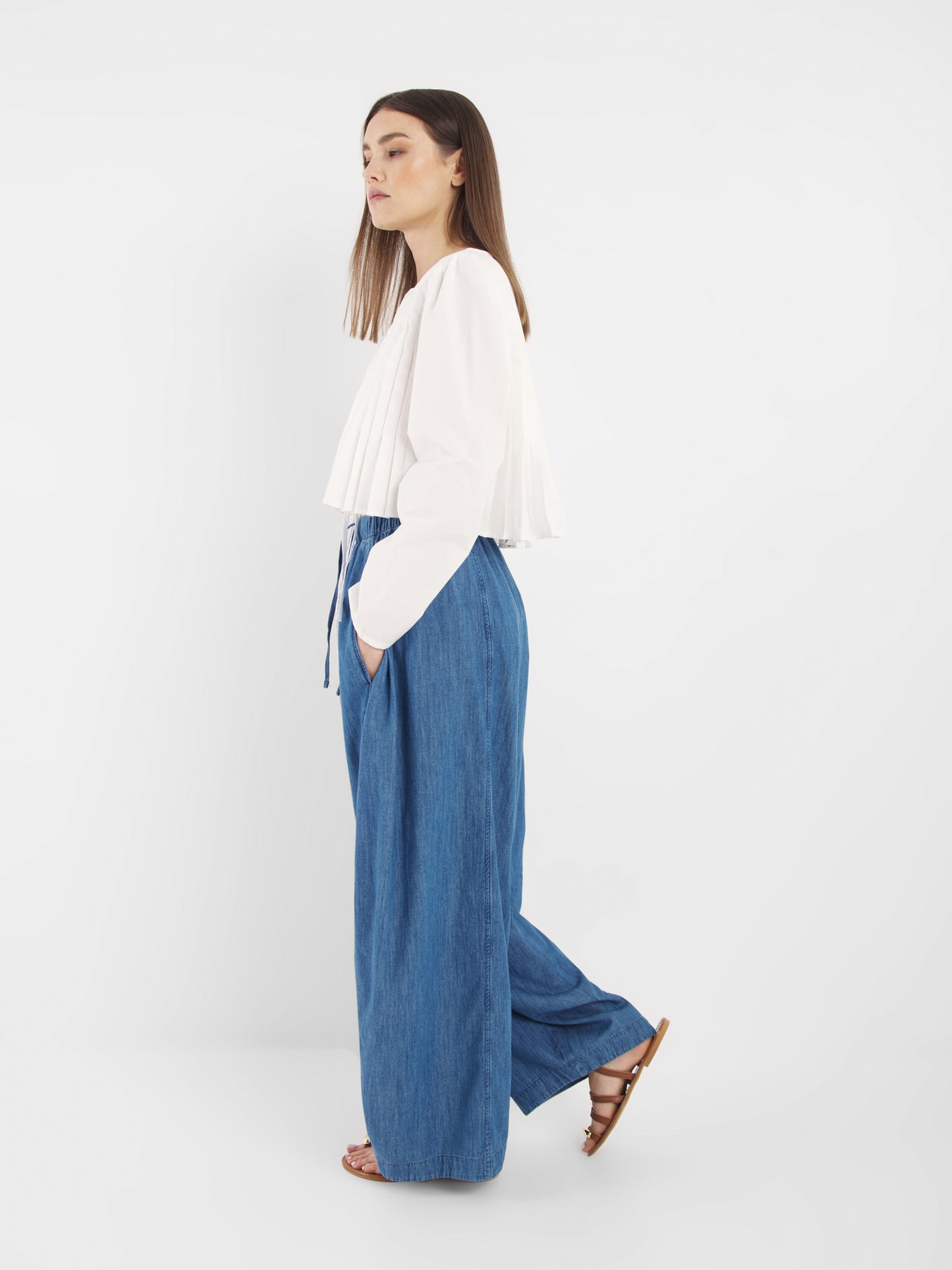 Eine Frau steht im Profil vor einem weißen Hintergrund. Sie trägt die Bluse Cleo White mit einer weit geschnittenen blauen Jeanshose und braunen Sandalen. Ihre Hände stecken in den Taschen und ihr glattes Haar fällt locker.