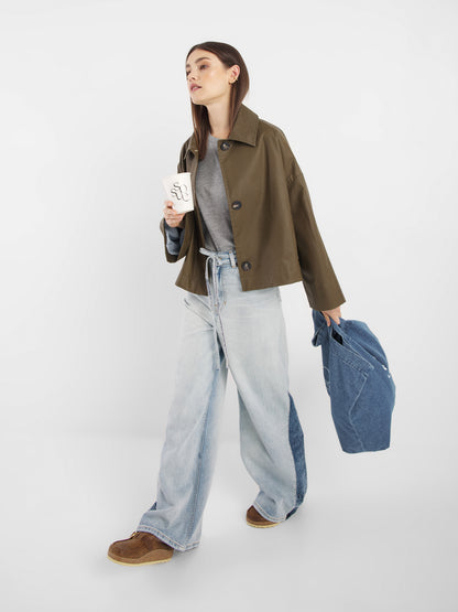 Eine Frau mit langen braunen Haaren trägt einen olivgrünen Dog-Walker-Mantel, ein graues T-Shirt, weit geschnittene hellblaue Jeans und braune Schuhe. Sie hält eine Jeansjacke und einen Kaffeebecher zum Mitnehmen in der Hand, während sie vor einem schlichten weißen Hintergrund geht.