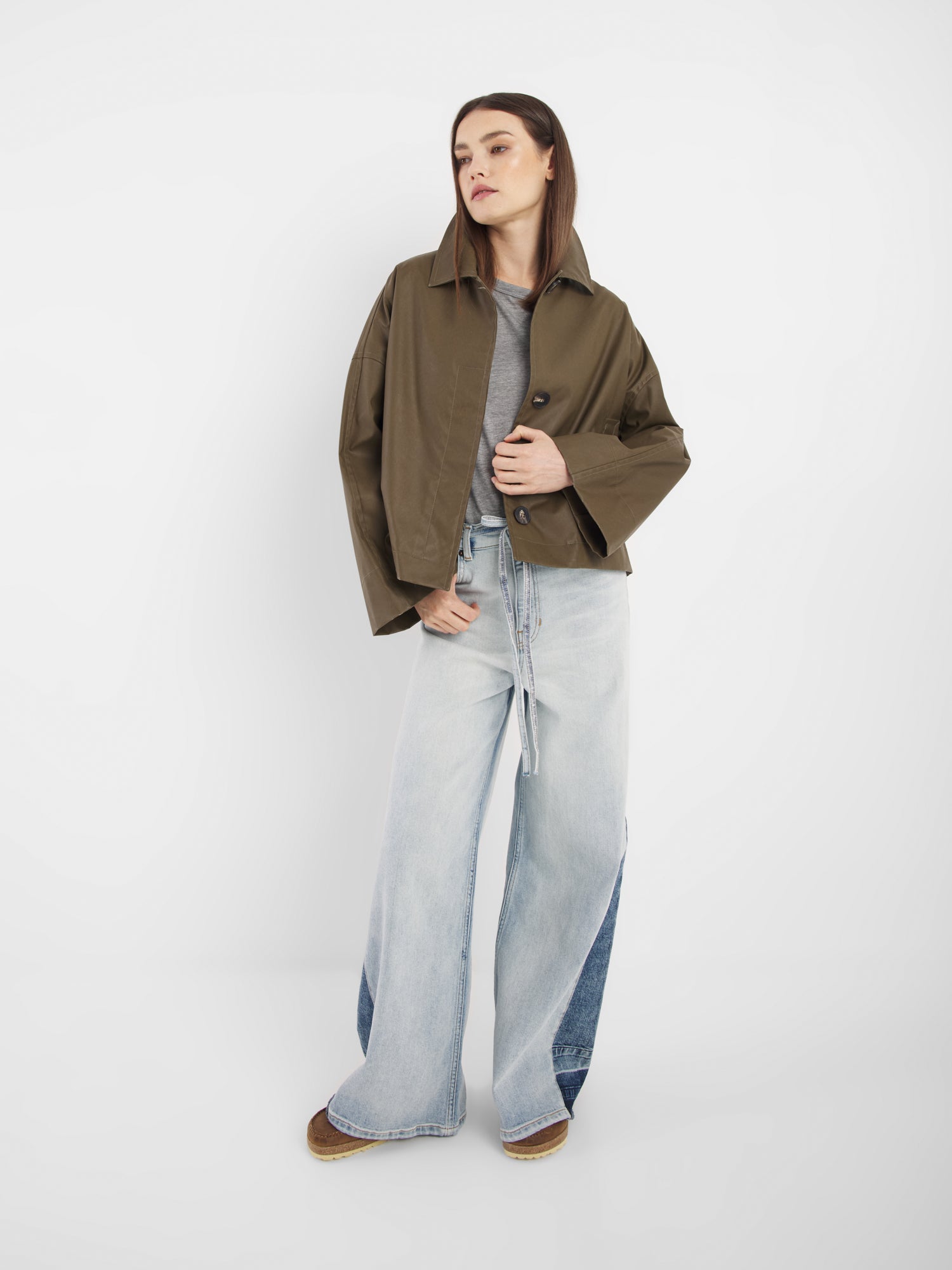 Eine Frau, die den Dog Walker Coat in Braun, eine moderne Übergangsjacke Damen, mit einem grauen T-Shirt, hellen Jeans mit weitem Bein und Denim-Patches und braunen Schuhen trägt, steht vor einem weißen Hintergrund und schaut zur Seite.