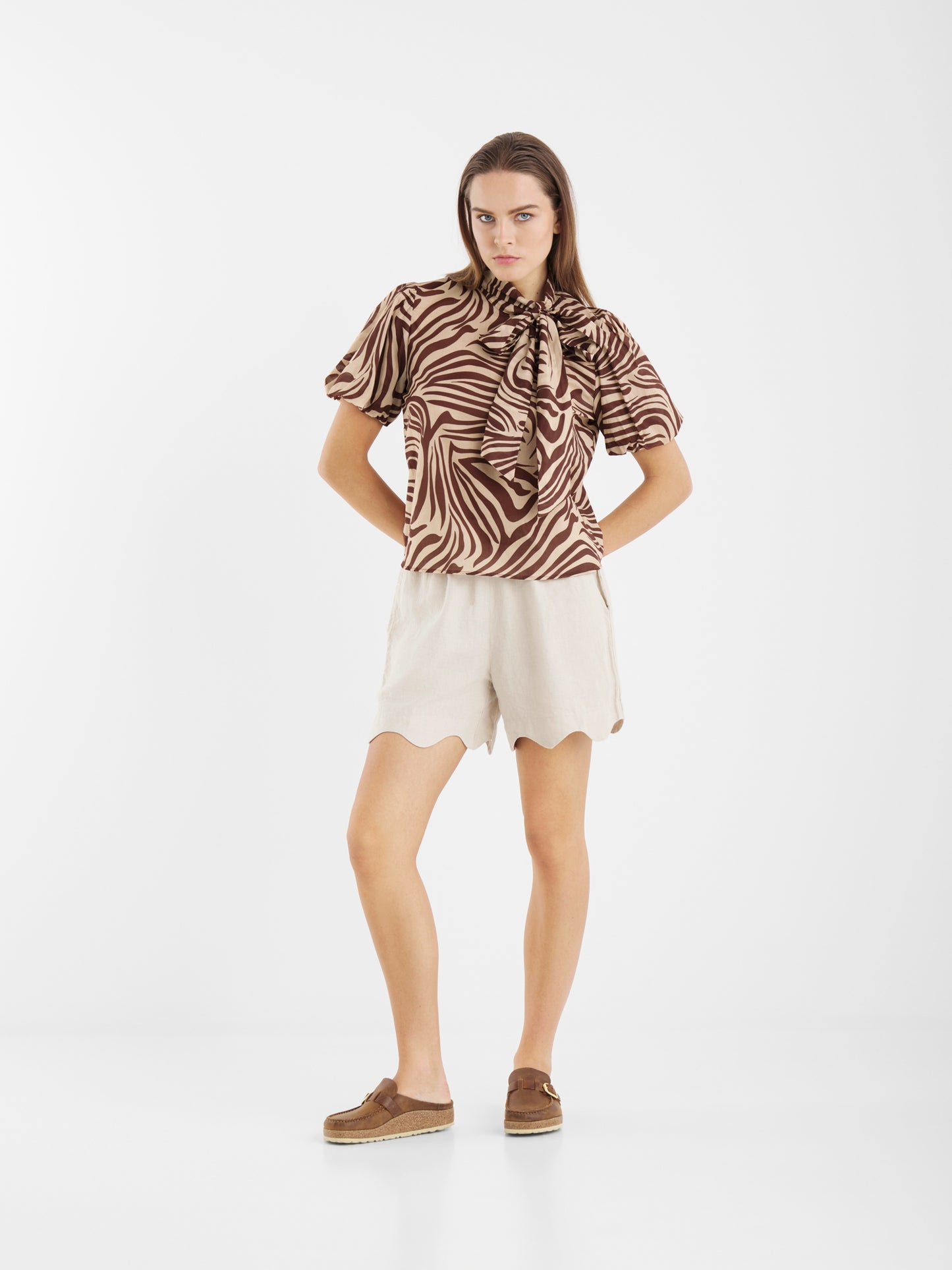 Eine Frau steht mit den Händen in den Hüften und trägt die Bluse Bowy Slim Zebra Brown aus leichtem Baumwoll-Voile, cremefarbene Shorts mit Wellenschliff und braune Slipper vor einem schlichten weißen Hintergrund.