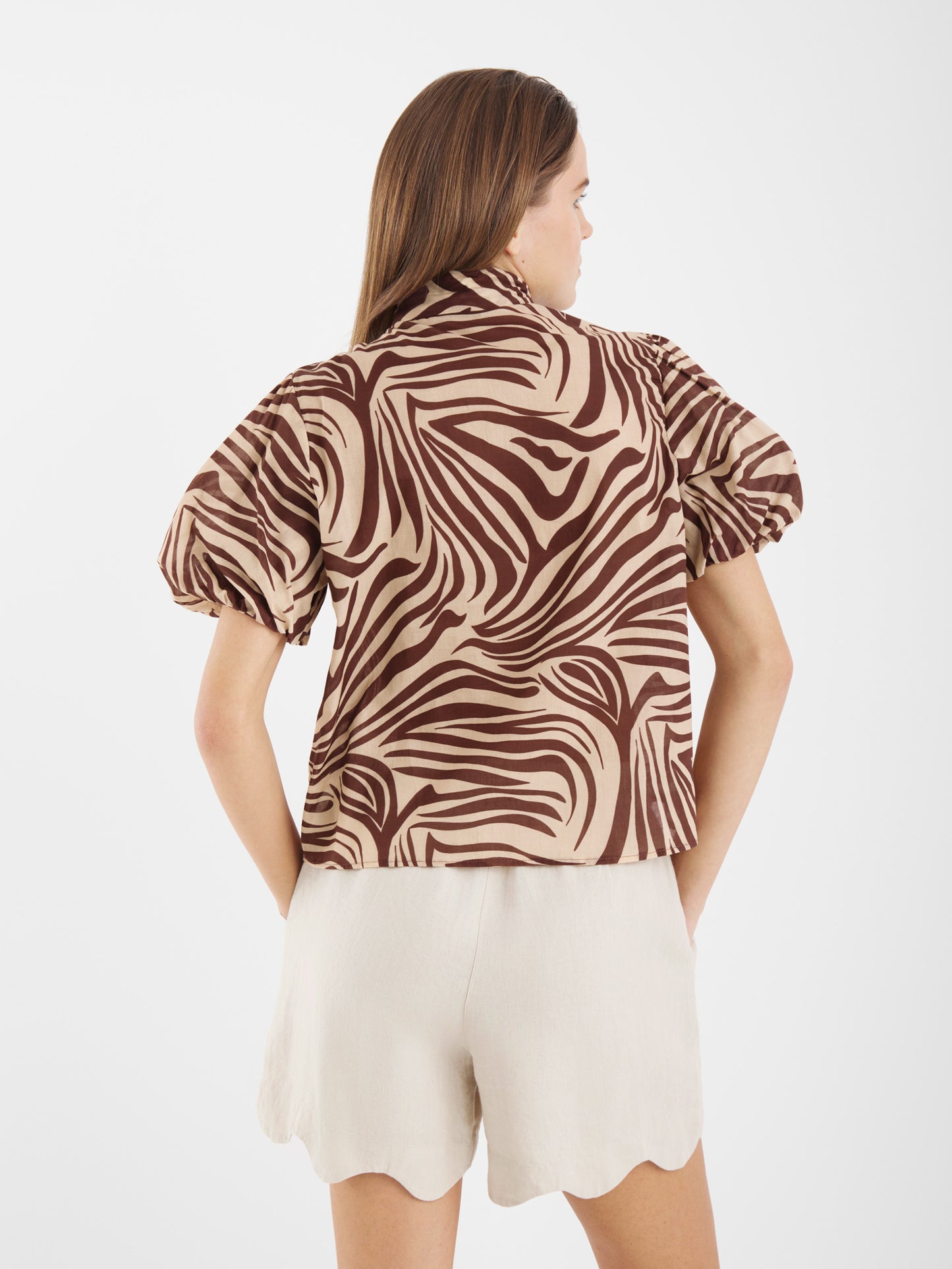 Eine Frau mit glattem braunem Haar ist von hinten zu sehen. Sie trägt die Bluse Bowy Slim Zebra Brown mit Puffärmeln mit Safariprint und hellbeigefarbenen Baumwoll-Voile-Shorts mit Wellenabschluss vor einem schlichten weißen Hintergrund.