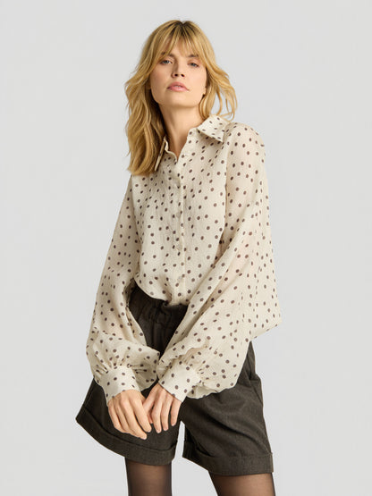 Eine blonde Frau trägt die Bluse „Antonia Polka Dots Ecru Brown“, eine cremefarbene Langarmbluse aus weichem Baumwollvoile, kombiniert mit dunklen Shorts mit hoher Taille vor einem hellgrauen Hintergrund.