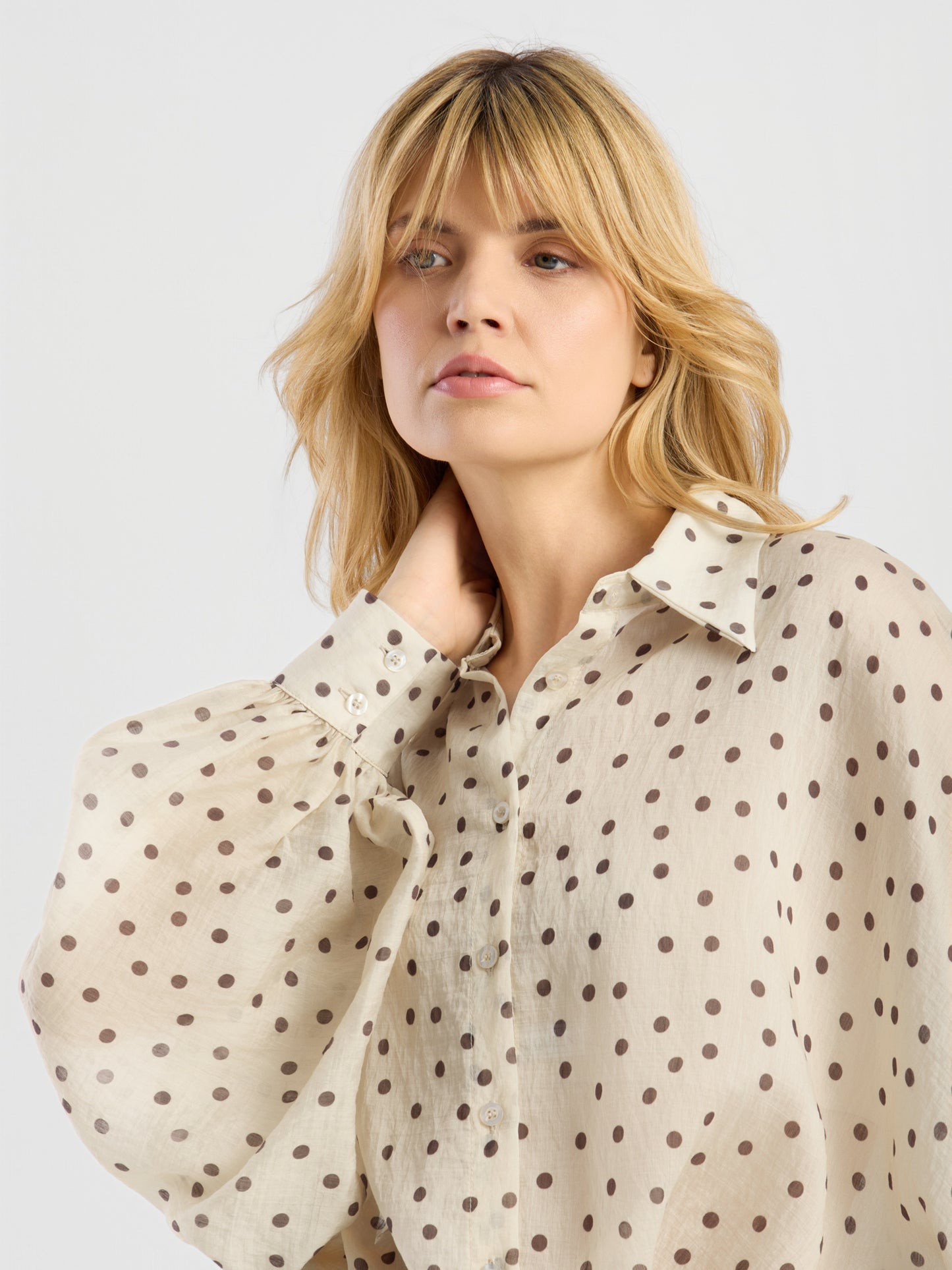 Eine Frau mit blonden Haaren trägt die Bluse „Antonia Polka Dots Ecru Brown“ mit braunen Punkten und Puffärmeln aus luftigem Baumwollvoile. Vor einem schlichten hellgrauen Hintergrund blickt sie mit ausdruckslosem Gesicht zur Seite, die Hand am Nacken.