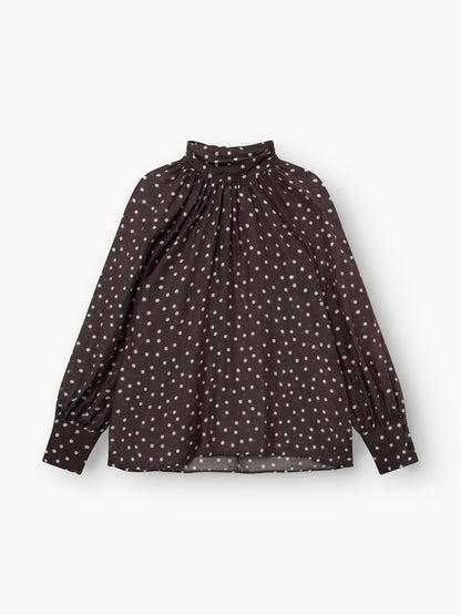 Die Bluse „Dotty Bow Brown Ecru“ ist eine langärmelige, dunkelbraune Bluse mit weißen Punkten, einem hoch gerafften Ausschnitt und Puffärmeln. Sie besteht aus weichem Baumwollvoile und wird flach auf einem weißen Hintergrund abgebildet.