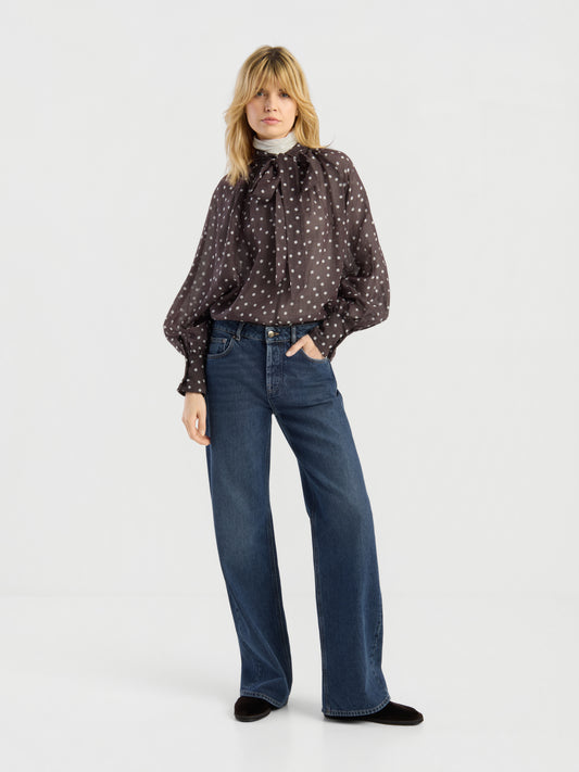 Eine Person trägt die schwarze, langärmelige „Dot Mood“-Bluse mit Polka-Dots und einer Schleife am Hals, dazu eine blaue Jeans mit weitem Bein und schwarze Schuhe; sie steht vor einem schlichten weißen Hintergrund.