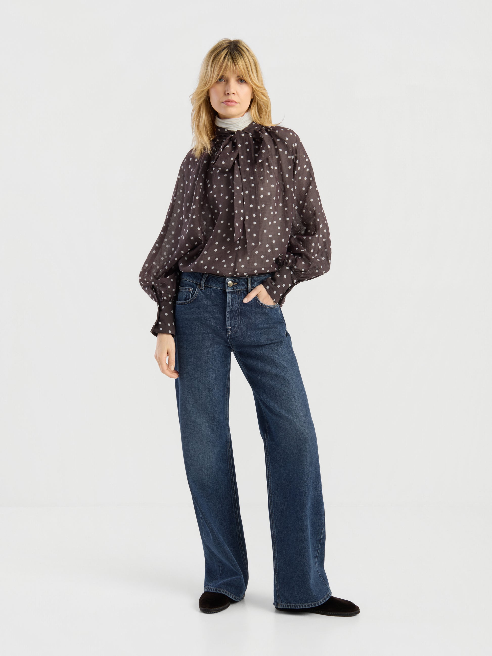 Eine Frau steht vor einem weißen Hintergrund und trägt die Bluse „Dotty Bow Brown Ecru“ mit einer großen Schleife am Hals, dazu eine blaue Jeans mit hoher Taille und weitem Bein sowie schwarze Schuhe.