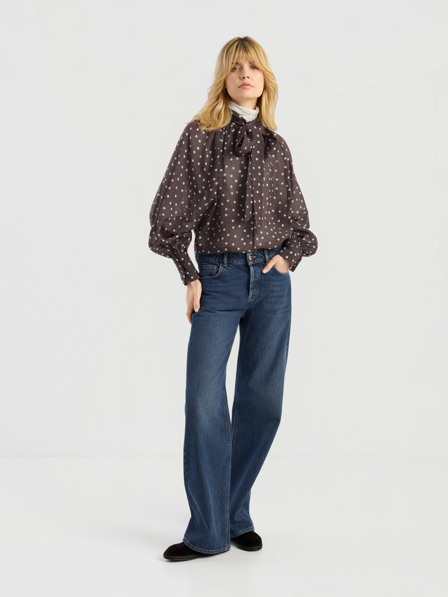Eine blonde Person trägt die Bluse „Dotty Bow Brown Ecru“ mit Schleifenkragen, eine blaue Jeans mit weitem Bein und schwarze Schuhe und steht vor einem schlichten weißen Hintergrund.
